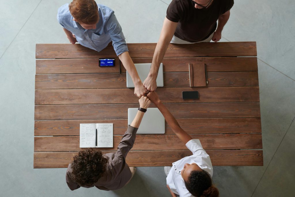 home A diverse group of professionals engaging in a teamwork celebration with a fist bump over a wooden table indoors.