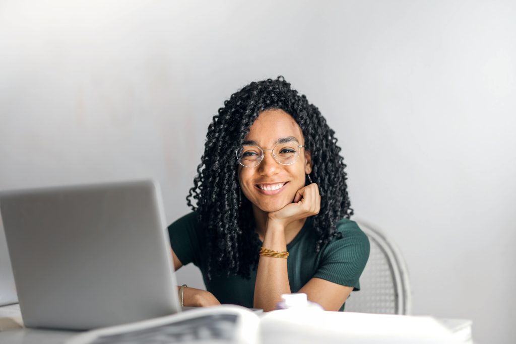 home Joyful businesswoman with curly hair smiling at camera while using laptop indoors.